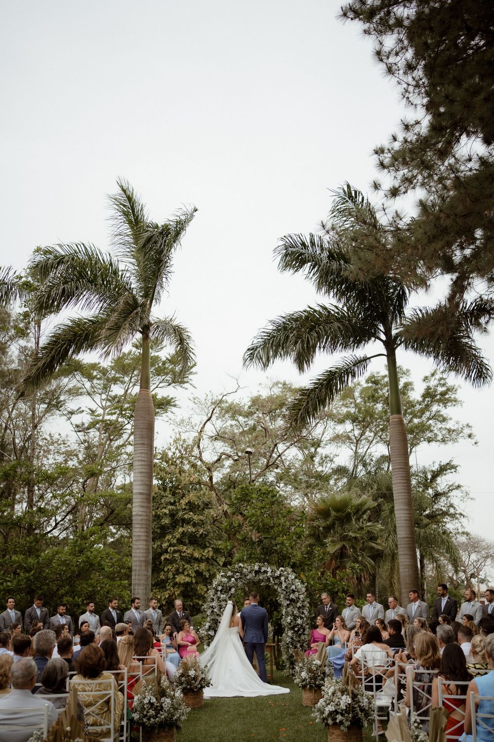 An outdoor wedding ceremony under tall palm trees with guests seated on wooden chairs and floral decorations.