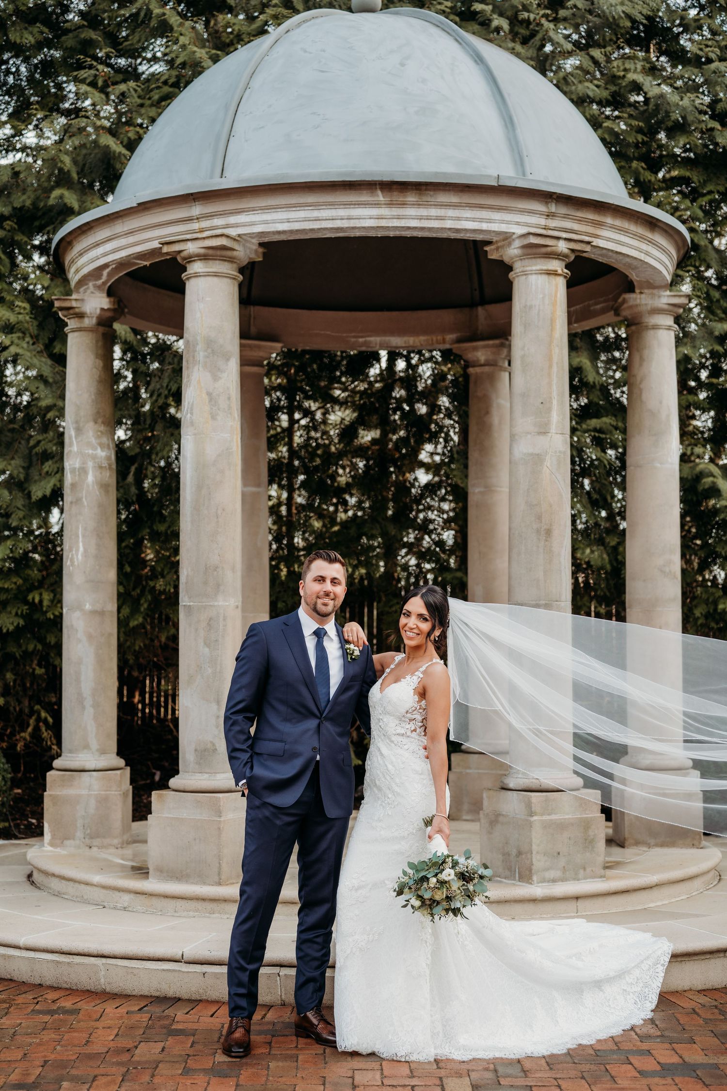 A couple stands under an elegant stone gazebo with a domed roof while a white veil flows in the breeze at Florentine Gardens