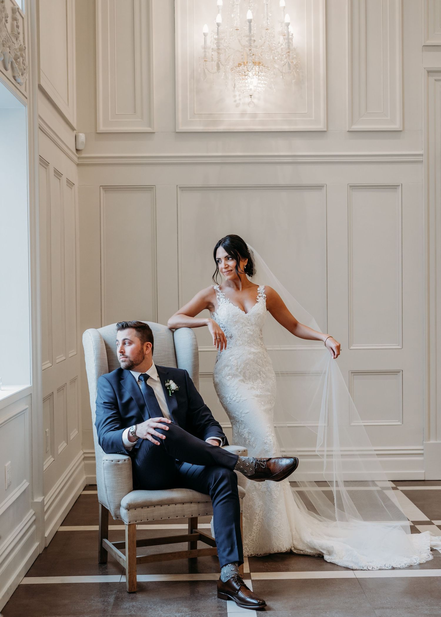 Elegant couple poses in formal wedding attire against white paneled wall with crystal chandelier in luxurious setting at Florentine Gardens.