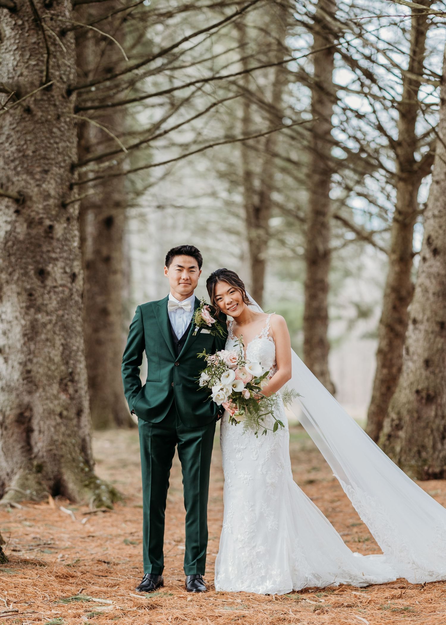 Newlyweds pose among tall pine trees in a romantic forest setting, with the bride holding a white floral bouquet at Perona Farms in Andover.