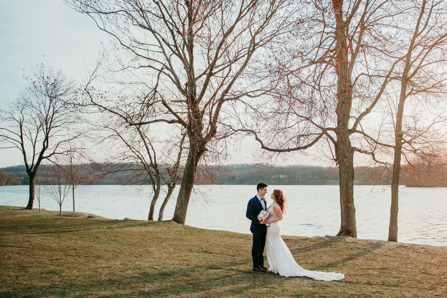 A romantic couple in wedding attire stands near a lake at sunset, surrounded by bare winter trees at Indian Trail Club.