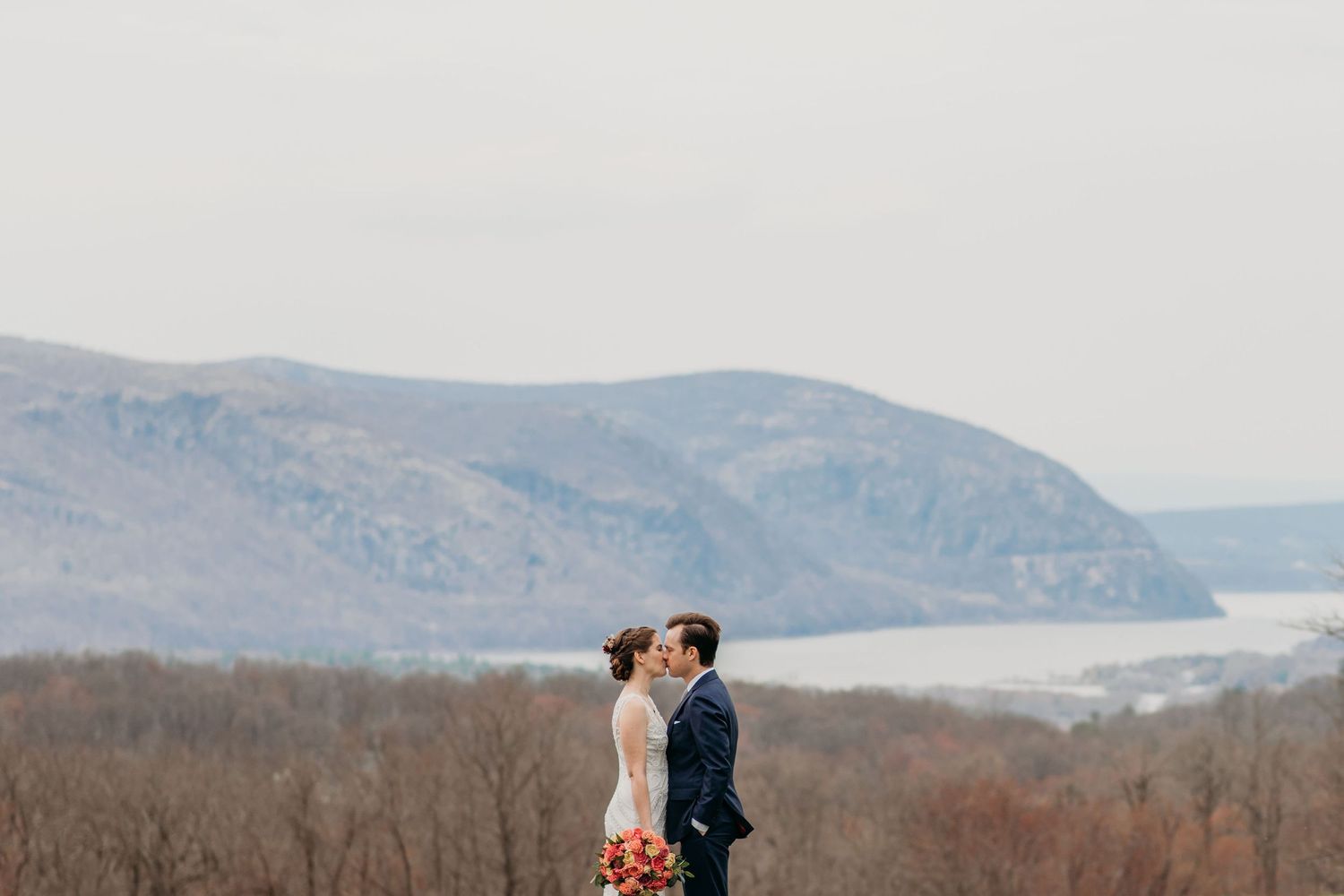 Couple stands together on a hillside overlooking a scenic mountain valley with misty blue peaks in the background.