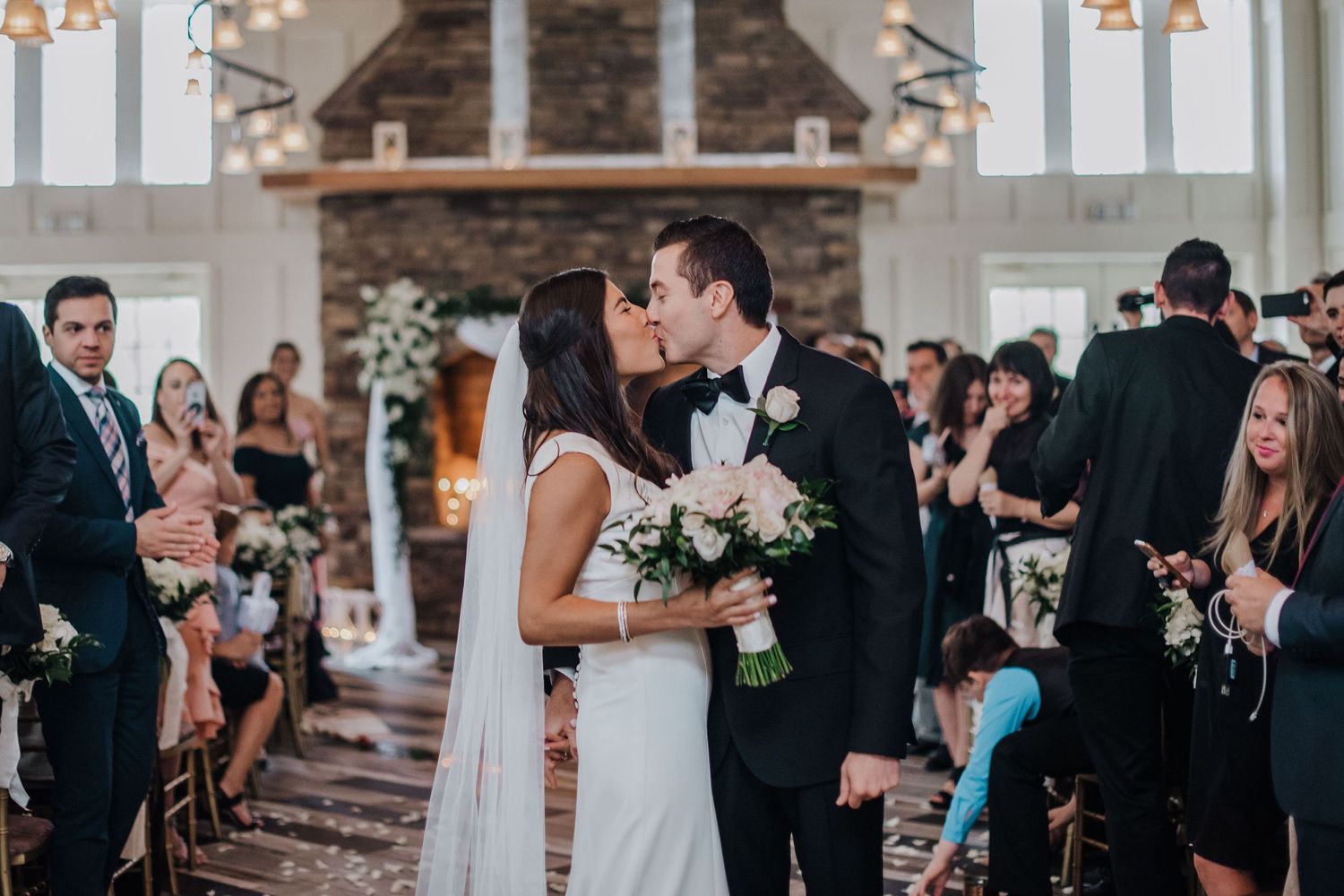 Newlyweds share a romantic kiss at altar during church ceremony while holding elegant white floral bouquet.