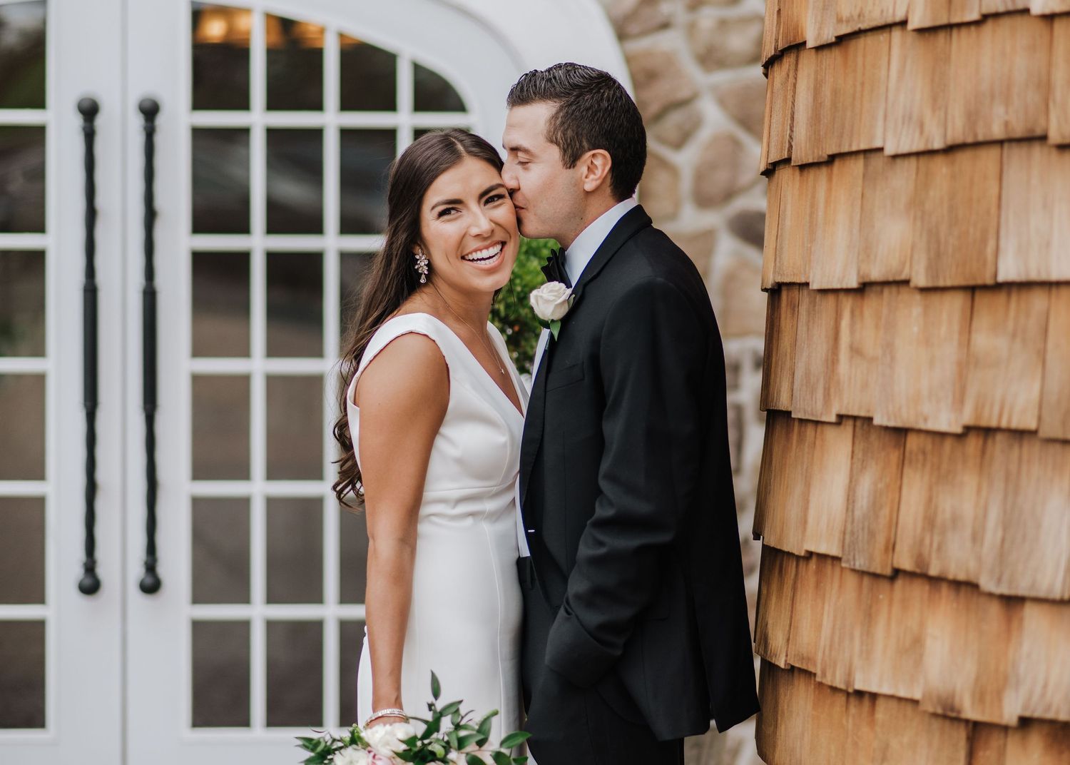 A happy couple embraces next to a cedar-shingled wall and white arched doorway in elegant wedding attire.