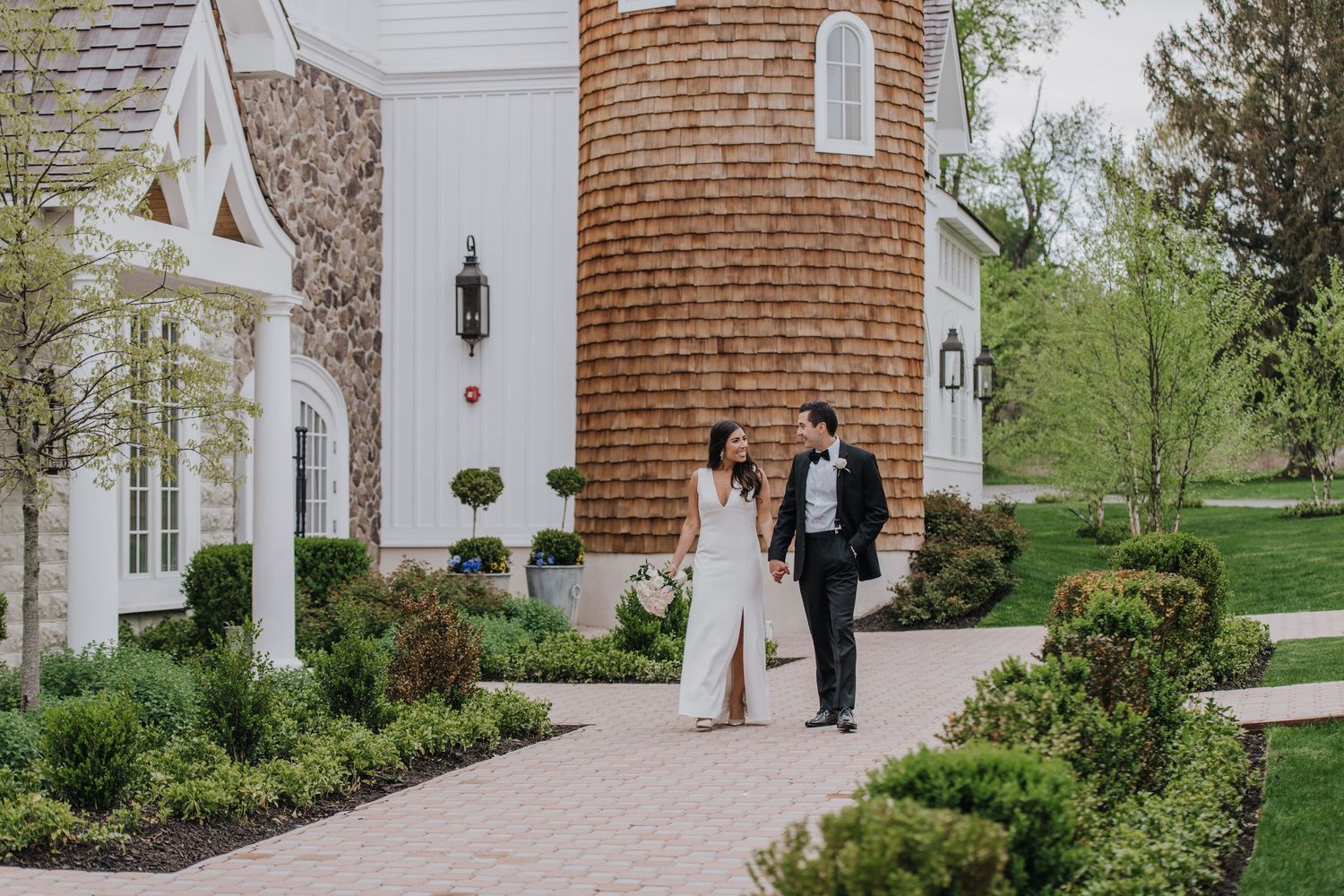 Elegant couple holding hands while walking along stone path by white chapel with rustic brick tower and garden landscaping at Ryland Inn.