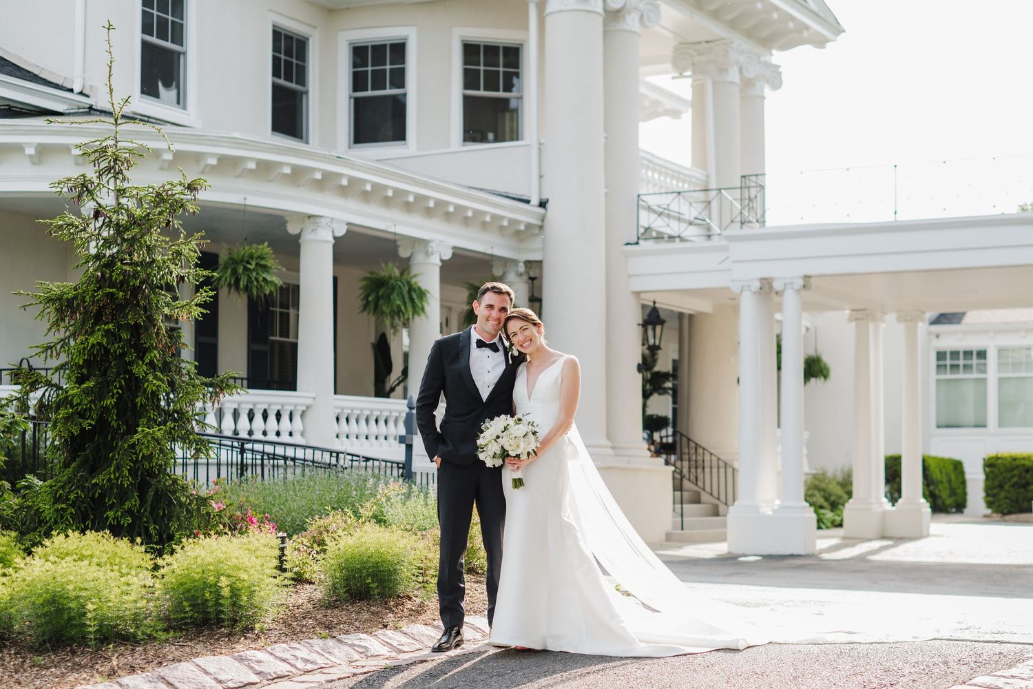 A happy couple in formal wedding attire stands in front of a grand white mansion with elegant columns and landscaping.