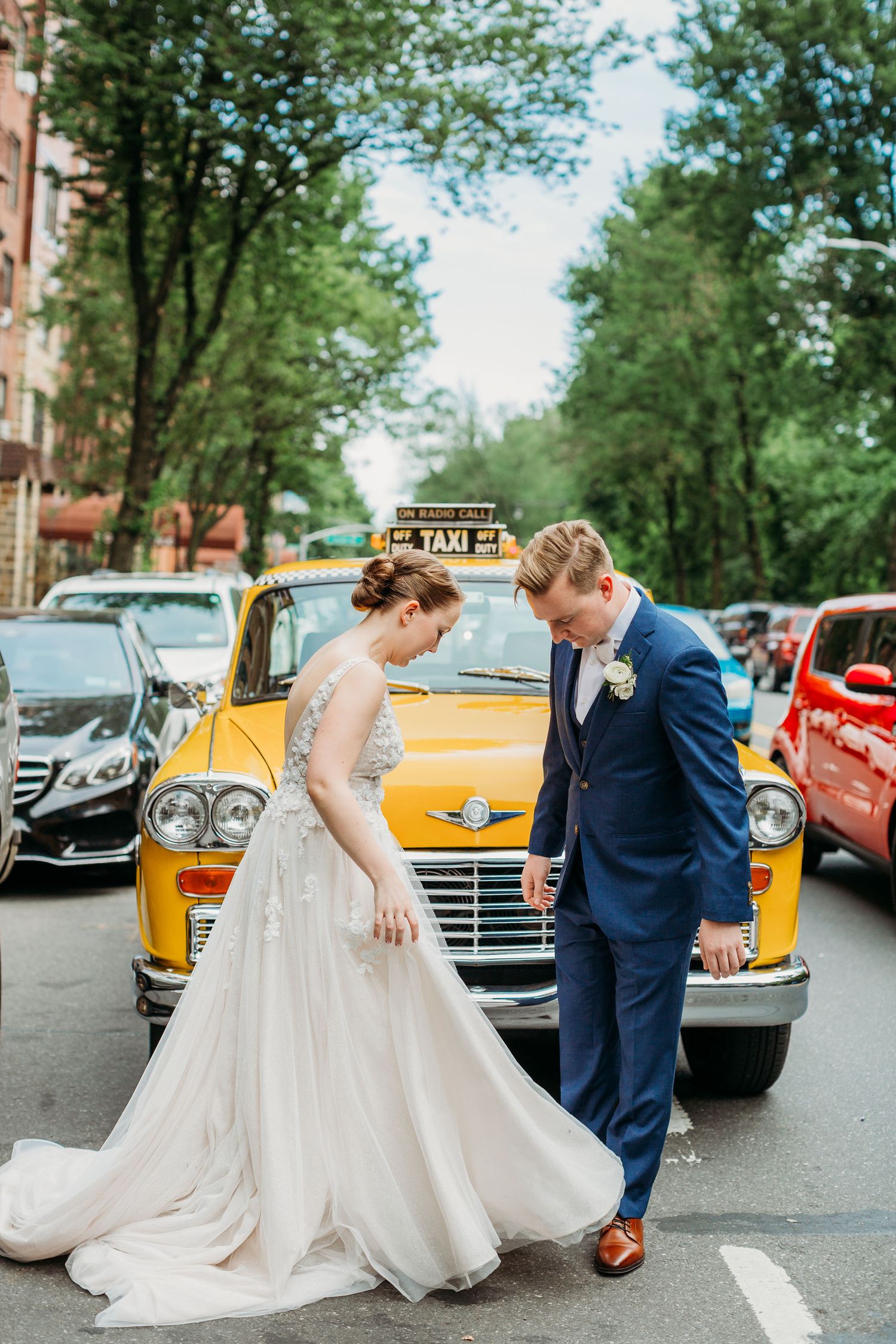 A couple poses in formal wedding attire in front of a vintage yellow taxi cab on a tree-lined city street.