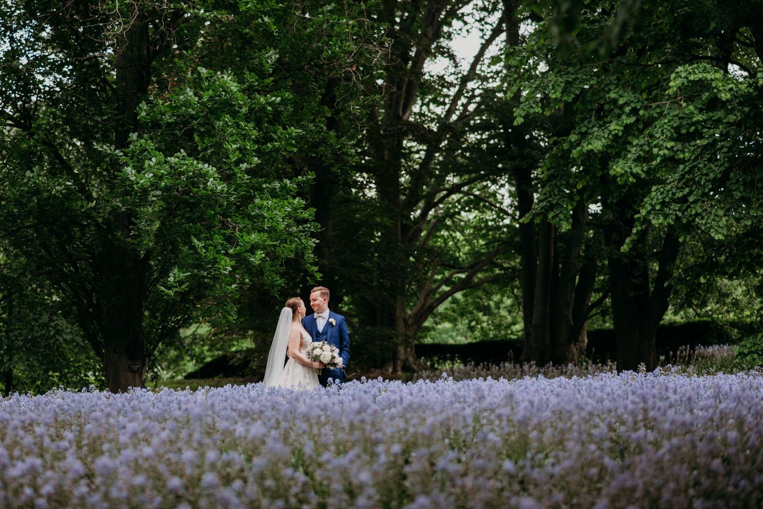 A couple stands together in a beautiful field of purple wildflowers surrounded by lush green trees at Brooklyn Botanical Garden.