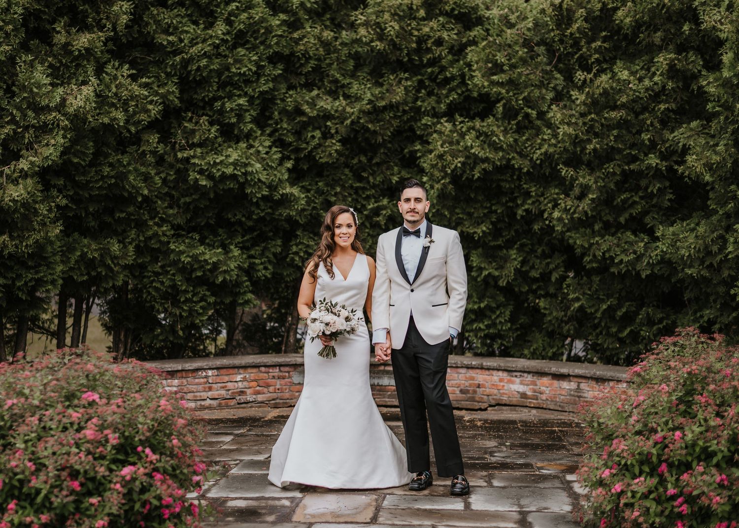 Couple in formal wedding attire poses together on stone path surrounded by lush gardens and pink flowers.
