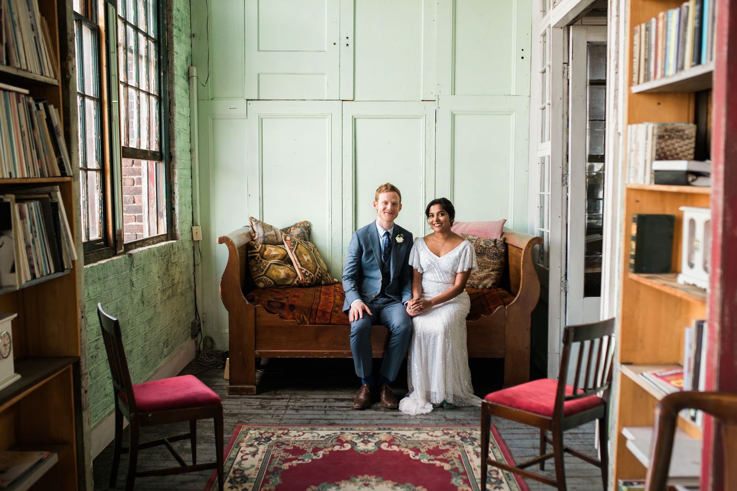 A couple sits on a vintage leather couch in a rustic room with bookshelves and large windows.