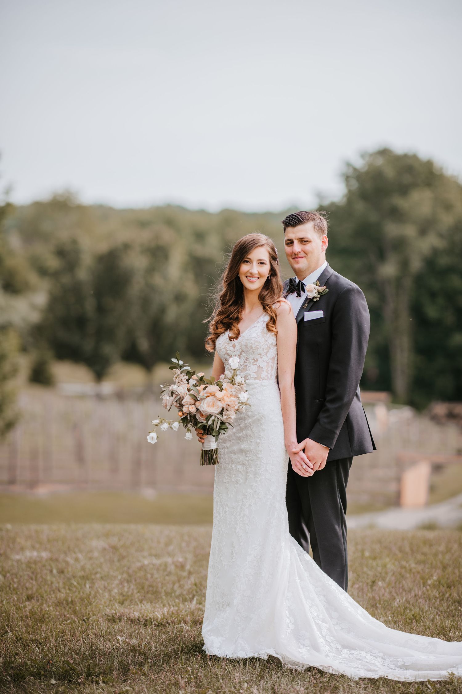 A couple in formal wedding attire stands together on grass with trees in the background while holding a floral bouquet.