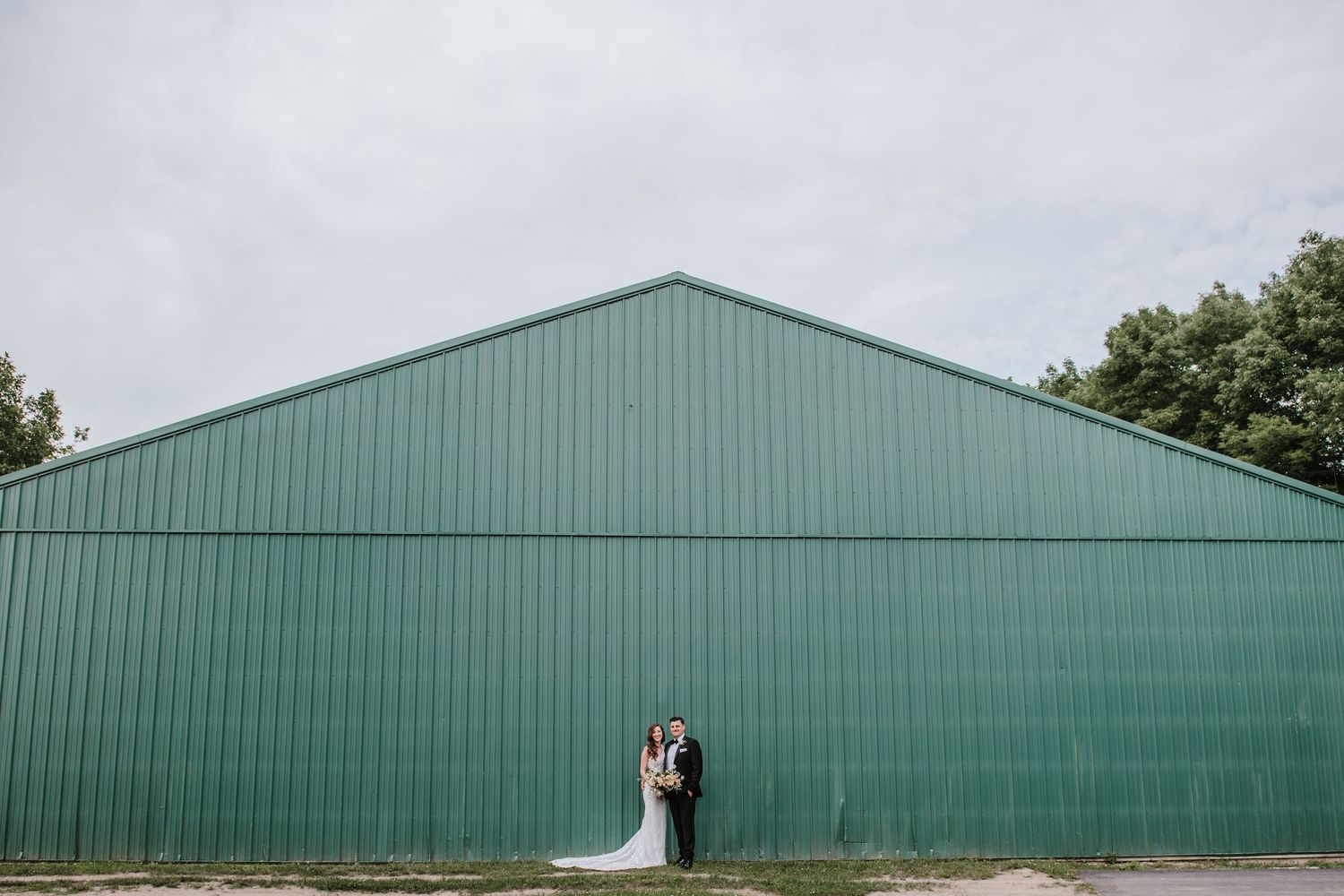 A couple stands in front of a large green industrial warehouse during a wedding photoshoot.