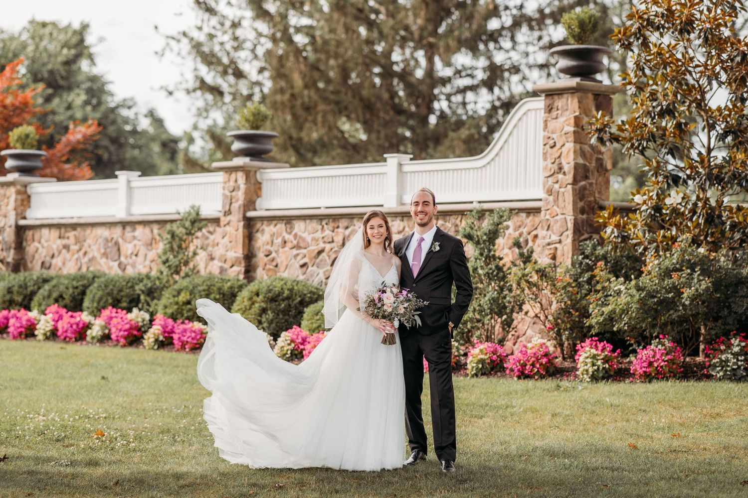 Couple poses on a garden lawn with pink azaleas and a stone wall backdrop during their elegant outdoor wedding ceremony.