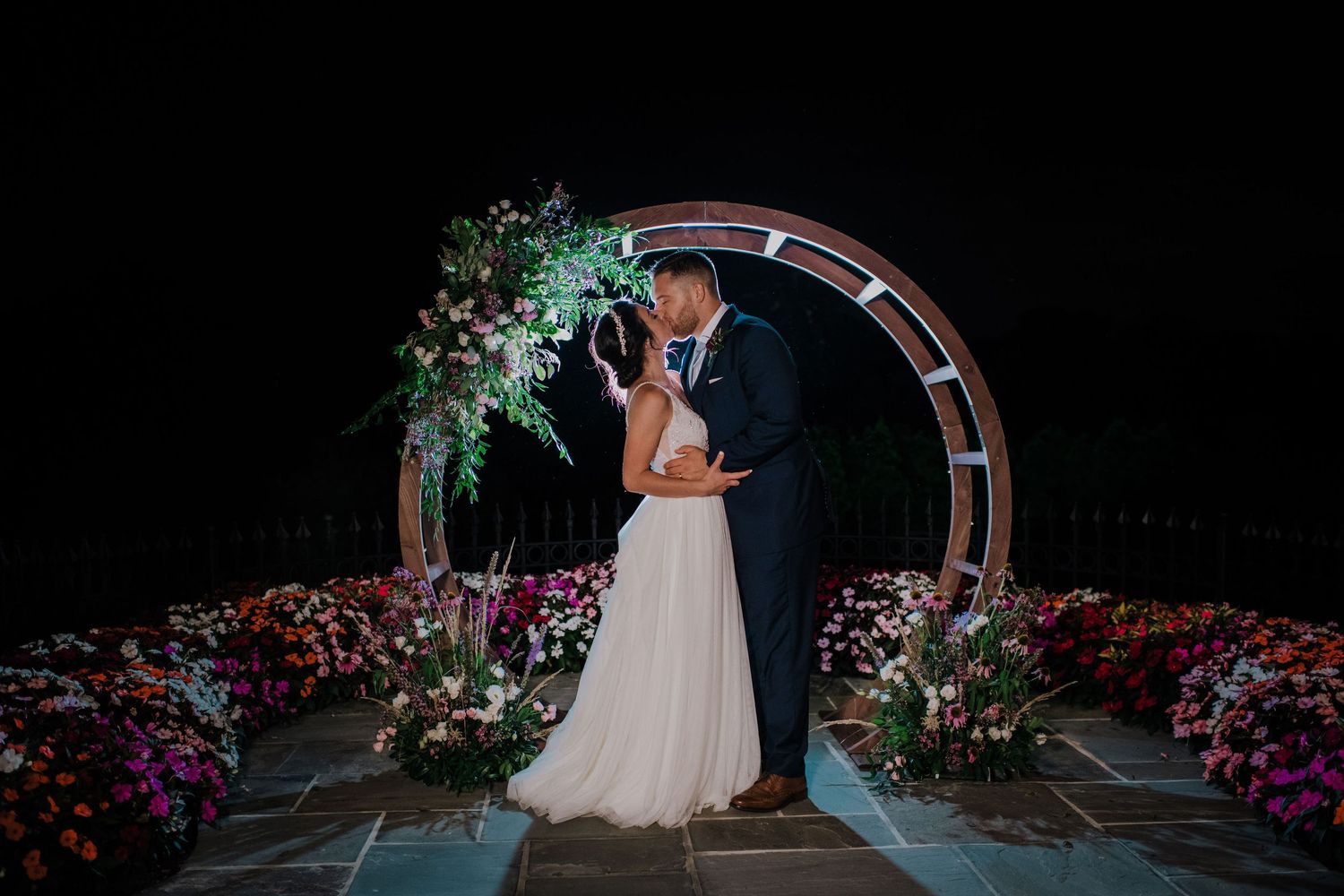 A couple embraces under an illuminated archway decorated with flowers and greenery during a nighttime wedding ceremony at the Park Savoy.
