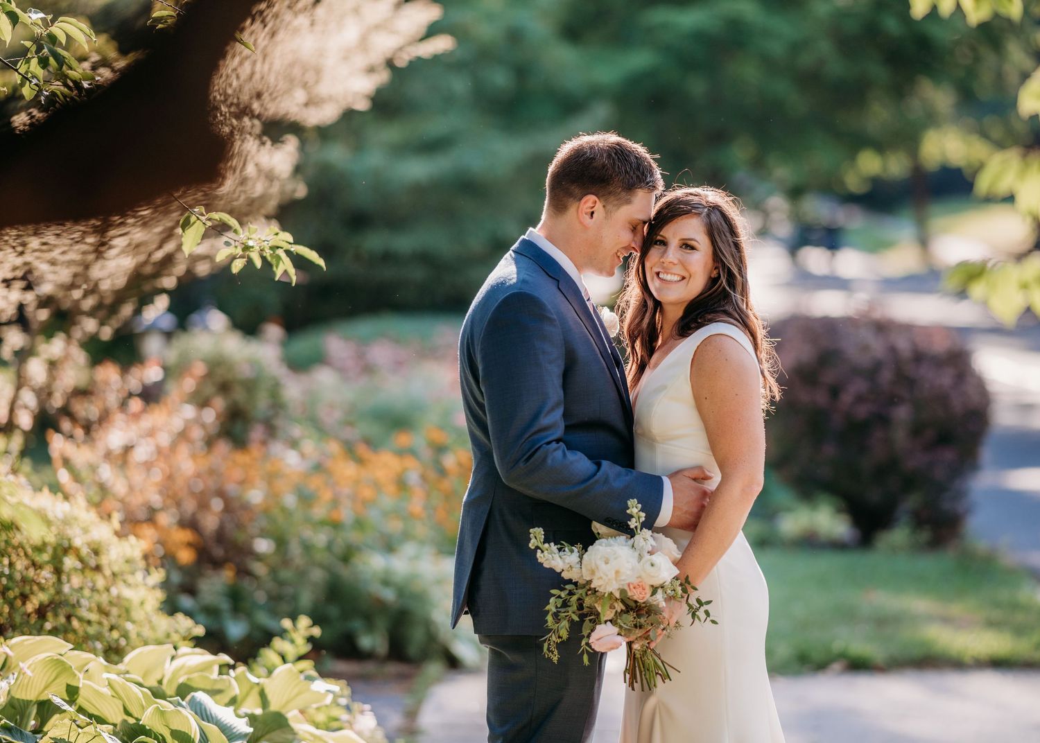 A couple shares an intimate moment in a garden setting with lush greenery and summer flowers in the background.
