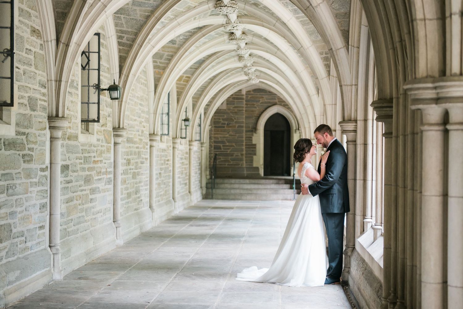 A couple in wedding attire embraces in a gothic stone archway corridor with vaulted ceilings & elegant architecture at Princeton University