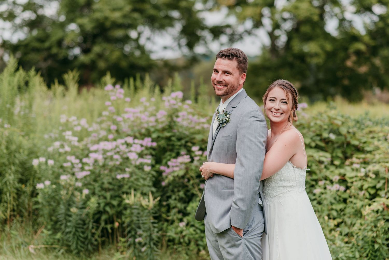 Happy newlyweds pose back-to-back in a lush garden setting with wildflowers and greenery on their summer wedding day at the Garrison in NY