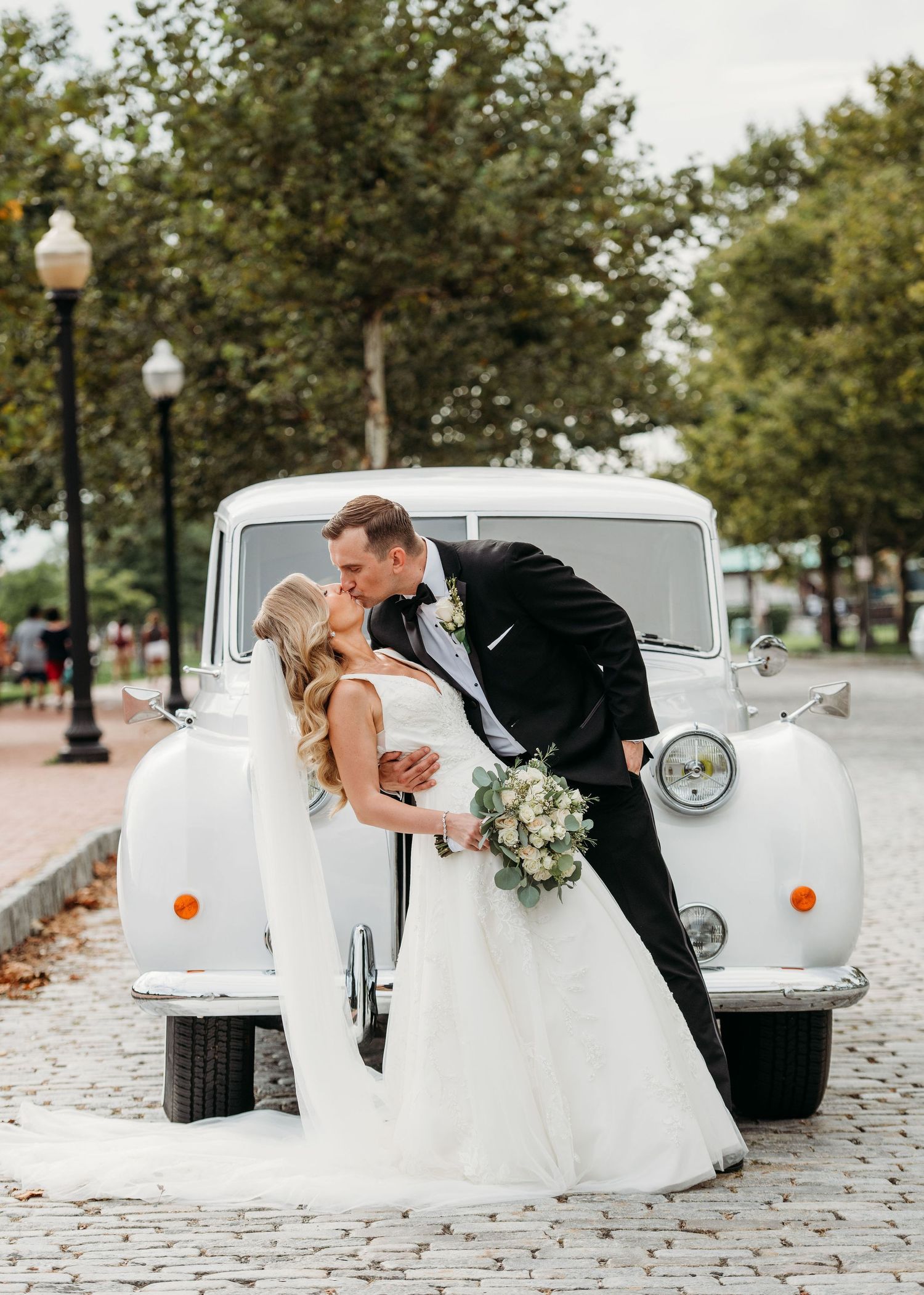 A couple poses romantically in front of a classic white vintage car on a brick-paved street with trees and lampposts.