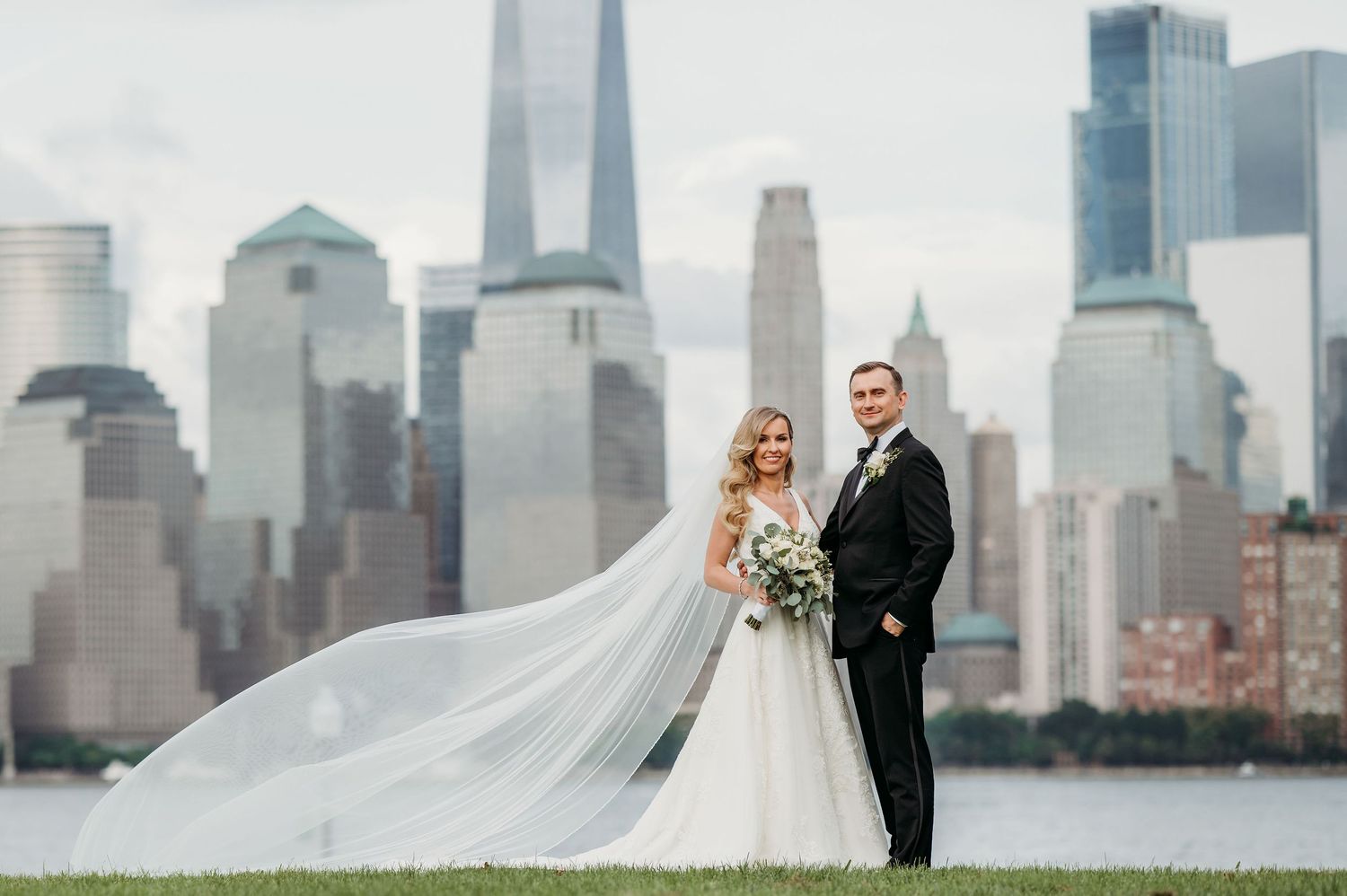 A couple poses for wedding photos with the dramatic New York City skyline and One World Trade Center in the background in Jersey City.
