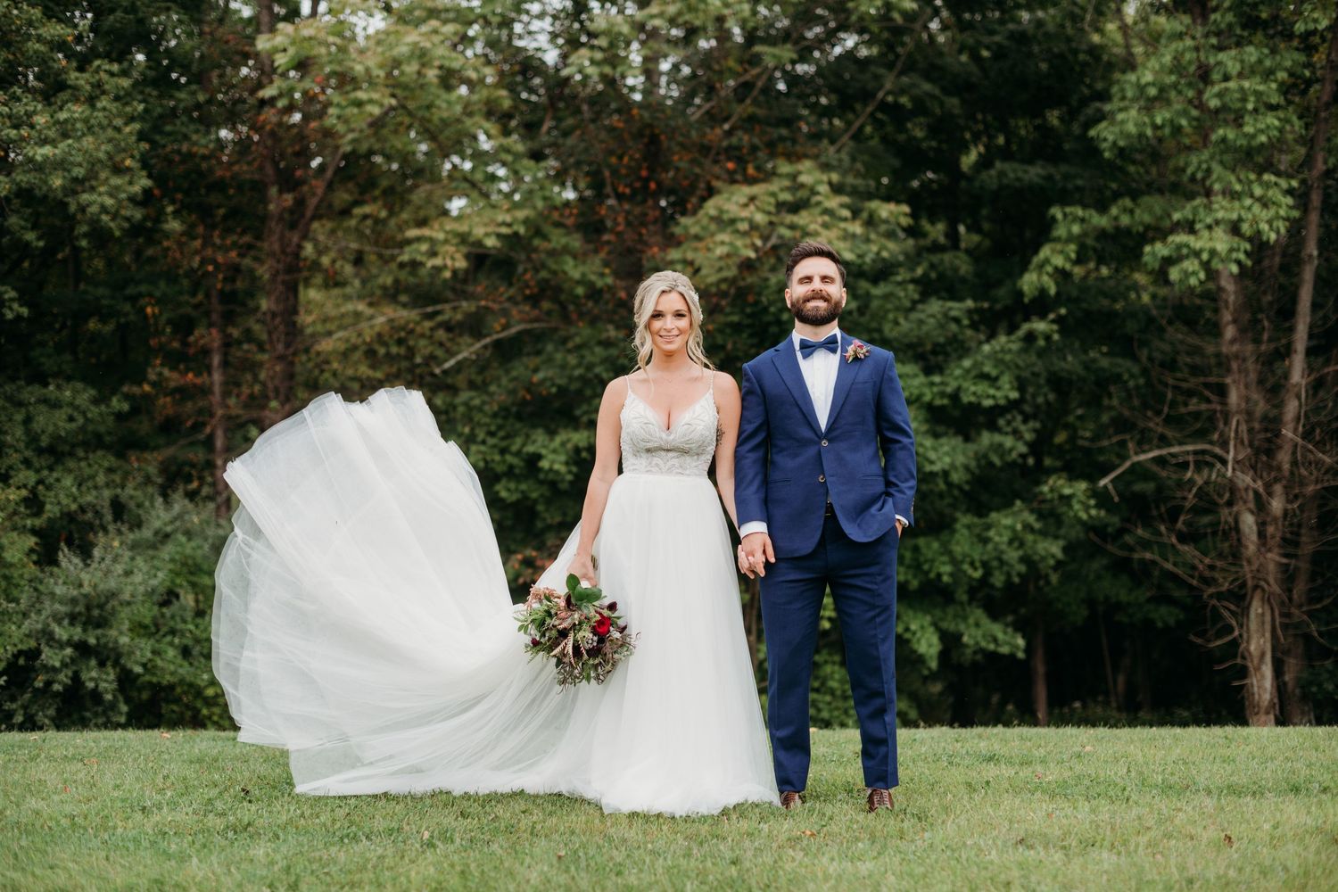 A couple in wedding attire stands together on a lush green lawn while the bride's white dress flows in the breeze.