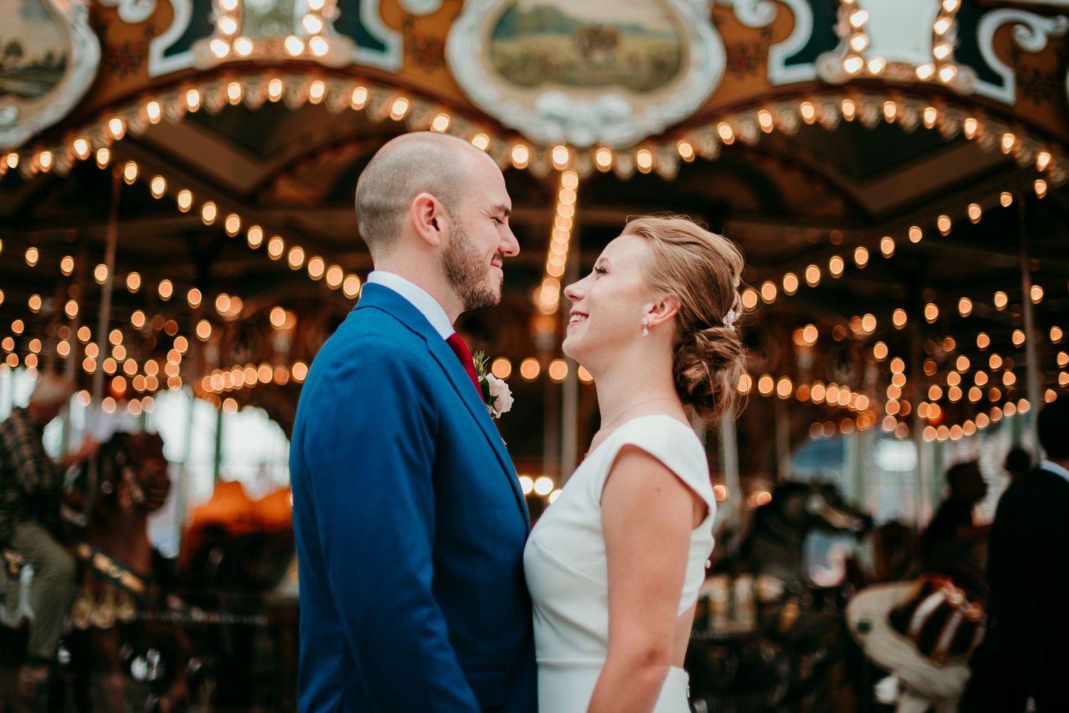 A couple shares an intimate moment in front of an illuminated vintage carousel with glowing lights at dusk at DUMBO in Brooklyn.