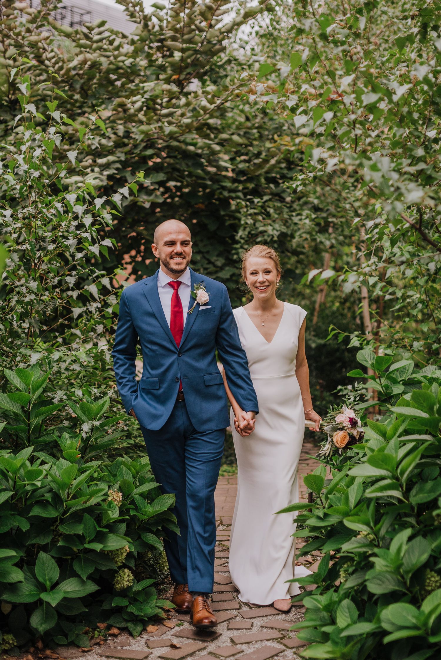 A bride and groom walk hand in hand along a garden path surrounded by lush greenery in Brooklyn.