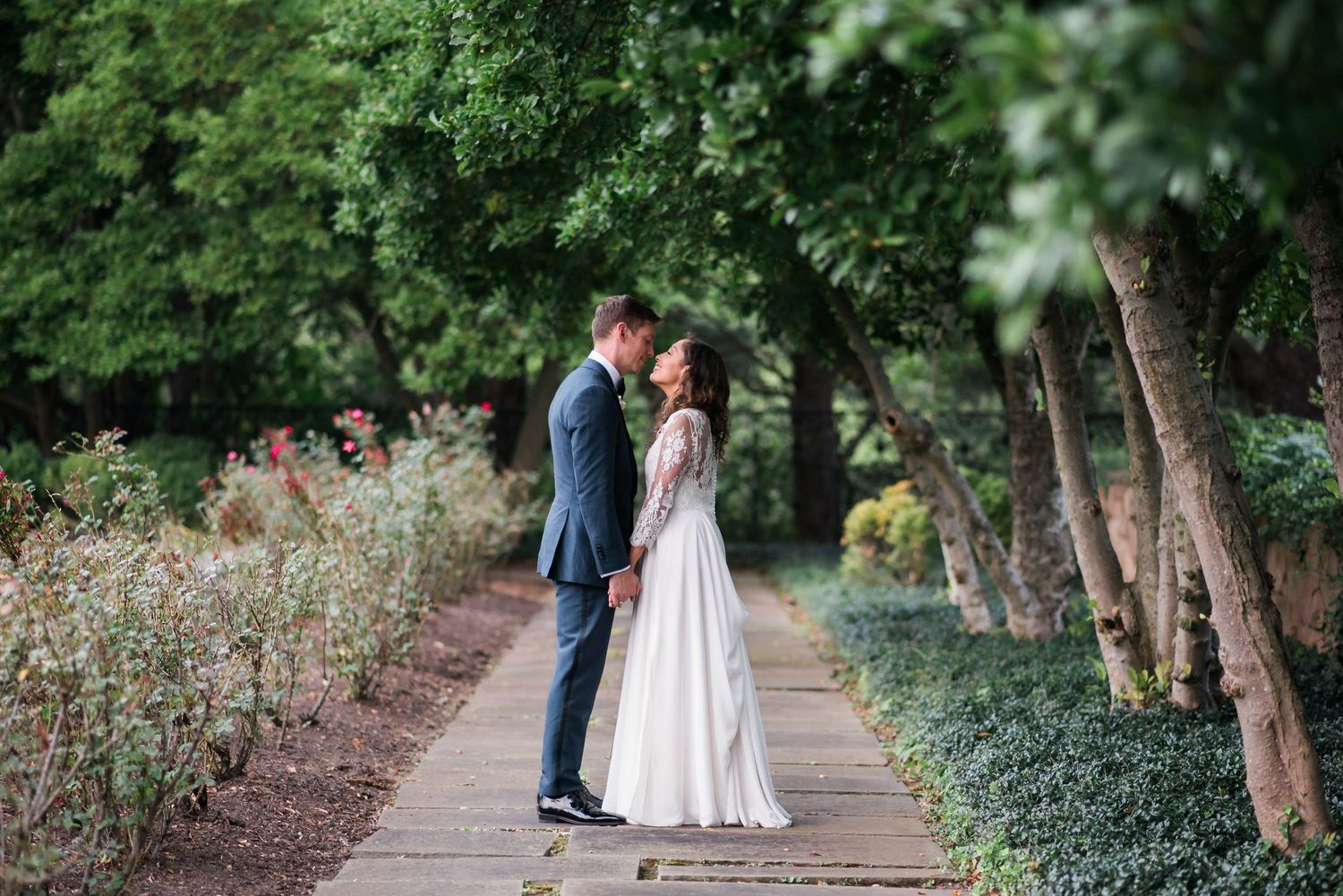 A couple in wedding attire embrace on a garden pathway lined with trees and flowering plants at Jasna Polana.