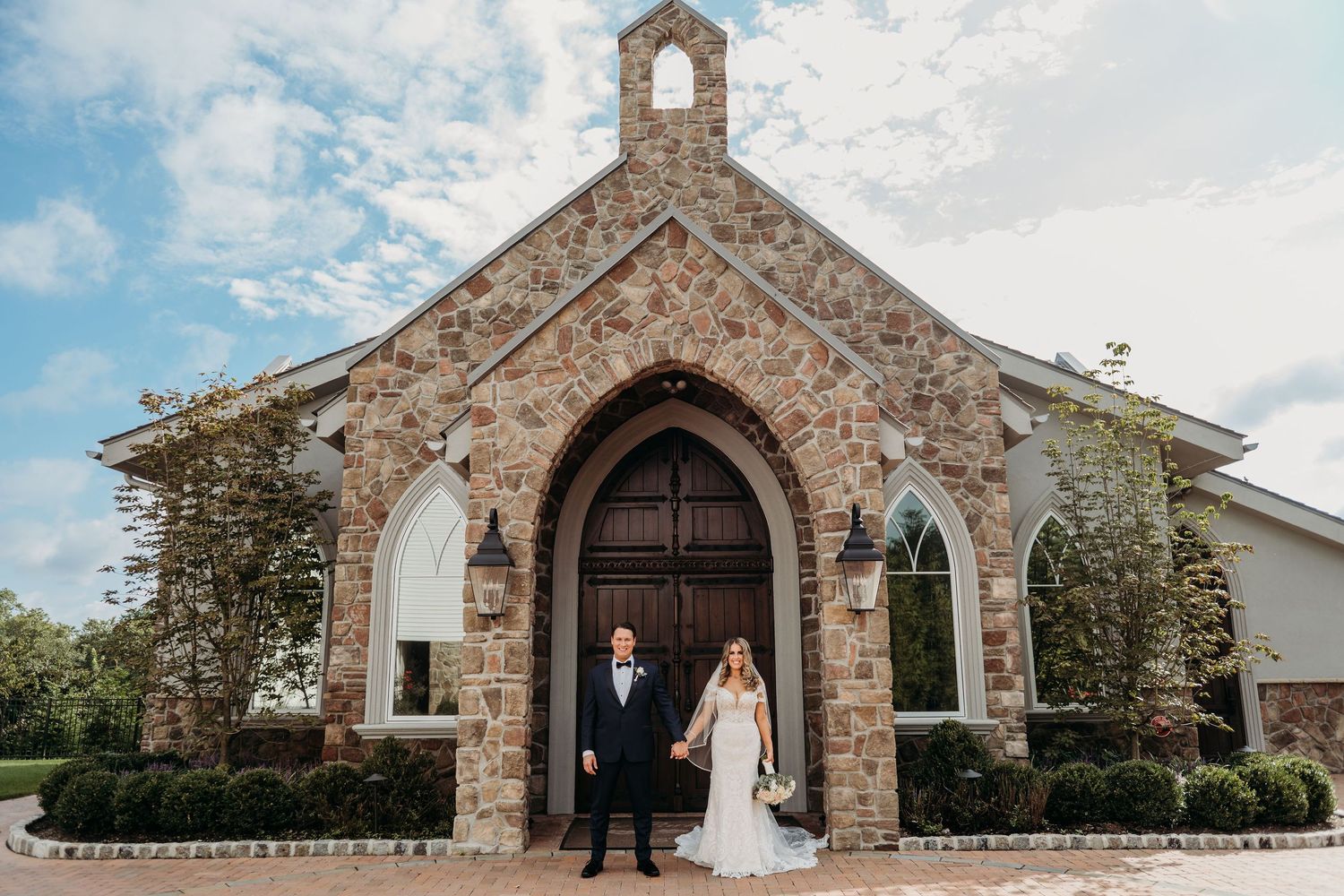 A charming stone chapel with Gothic arched doorway and white trim serves as backdrop for a wedding day photo.