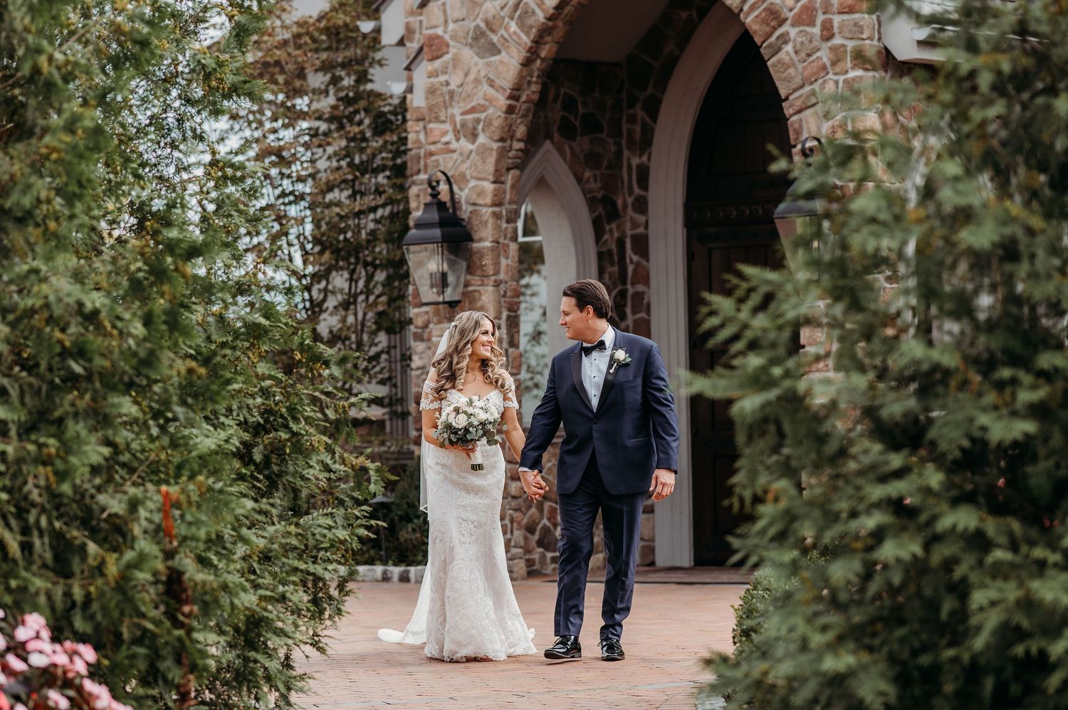 A bride and groom walk together outside a stone archway decorated with greenery and flowers.