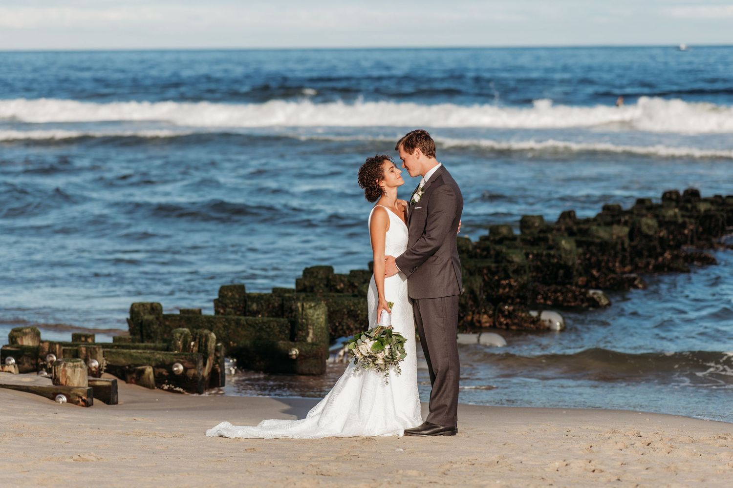 A couple shares a romantic moment on a sandy beach with waves crashing against a stone jetty in the background at Spring Lake.