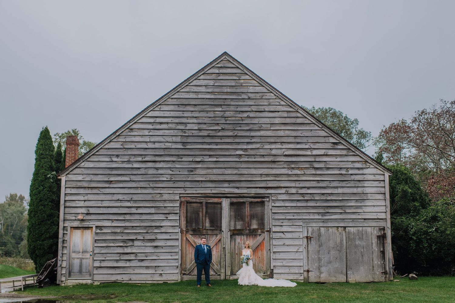 A rustic weathered wooden barn with faded gray siding stands against an overcast sky and green trees.