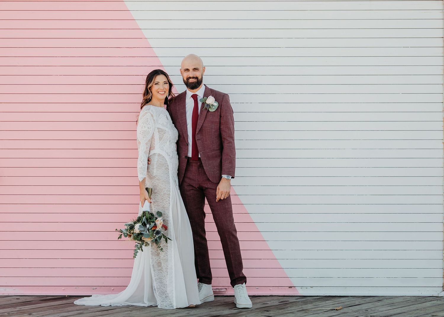 A couple in wedding attire poses against a pink and white geometrically painted wall with bridal bouquet in Asbury park at the Crepe Shop.