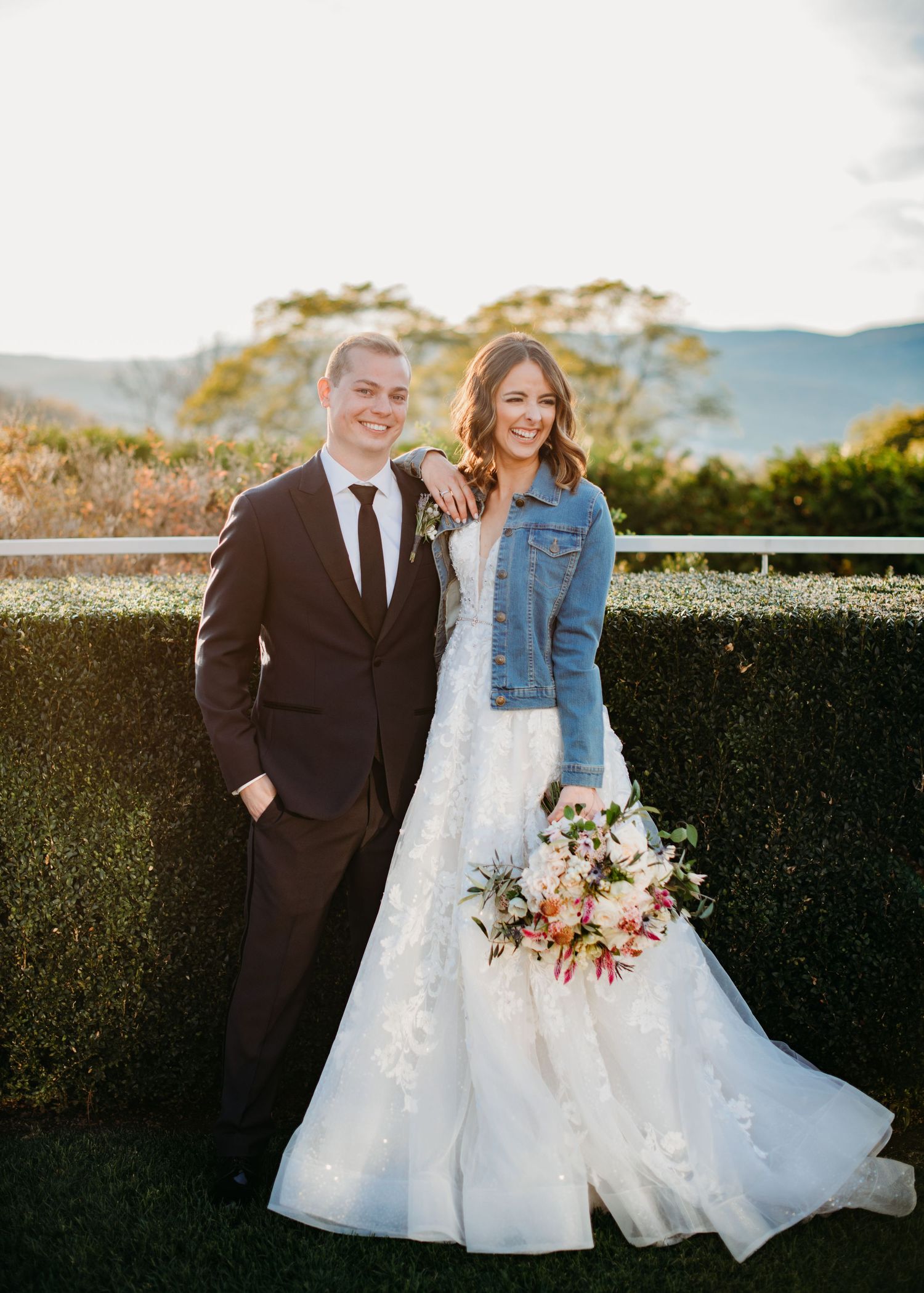 A couple poses outdoors at a garden venue, with the bride wearing a white gown and denim jacket holding a pink bouquet at the Garrison.