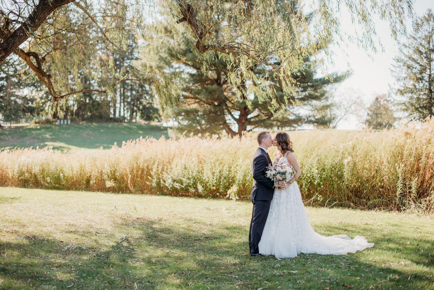 Two figures stand together in a sunny field with tall grass and evergreen trees in the background on a bright autumn day.