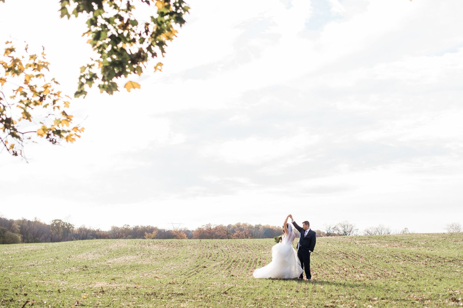 Newlyweds walking together in a scenic autumn field under a bright overcast sky with fall foliage in the distance at Hotel du Village.