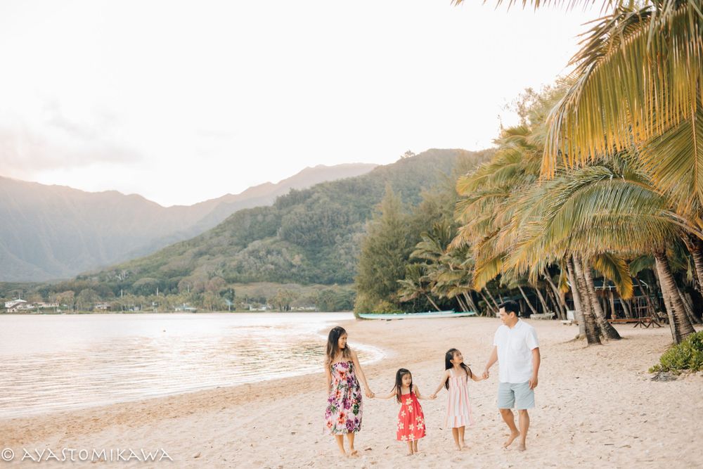 A family walking along a palm-lined beach with mountains in the background at sunset.