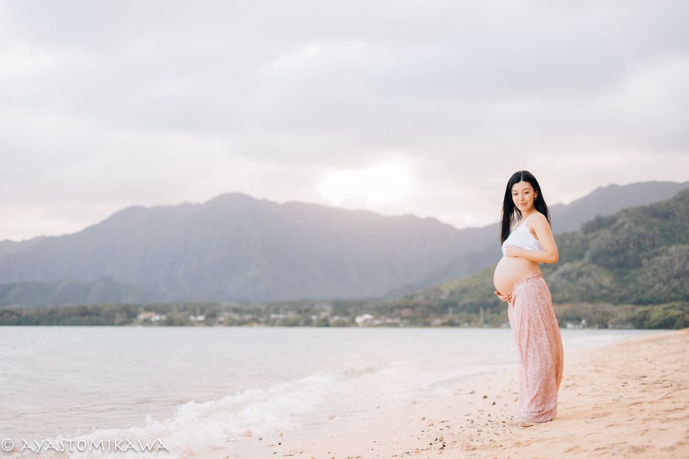 A woman in flowing dress poses on a beach with mountains and sunrise in the background.