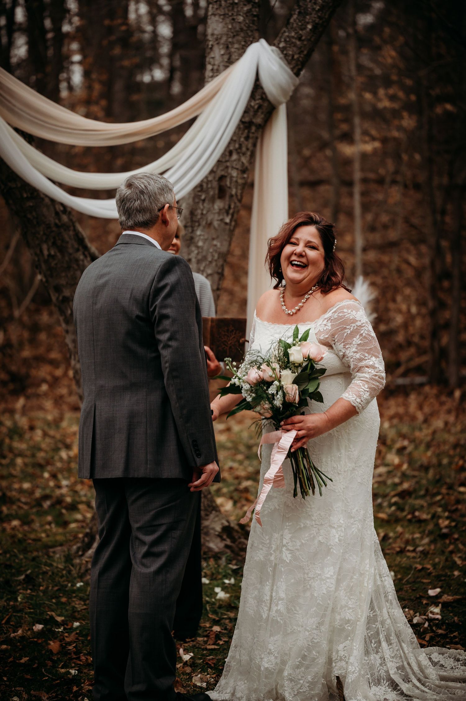 Smiling bride in lace gown holding bouquet during outdoor autumn wedding ceremony.