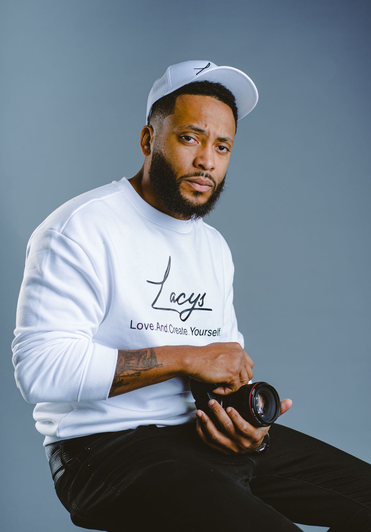 Professional photographer wearing white cap and sweater while holding camera against grey background.