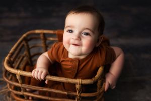 Baby sitting in wooden basket against dark backdrop in brown tones.