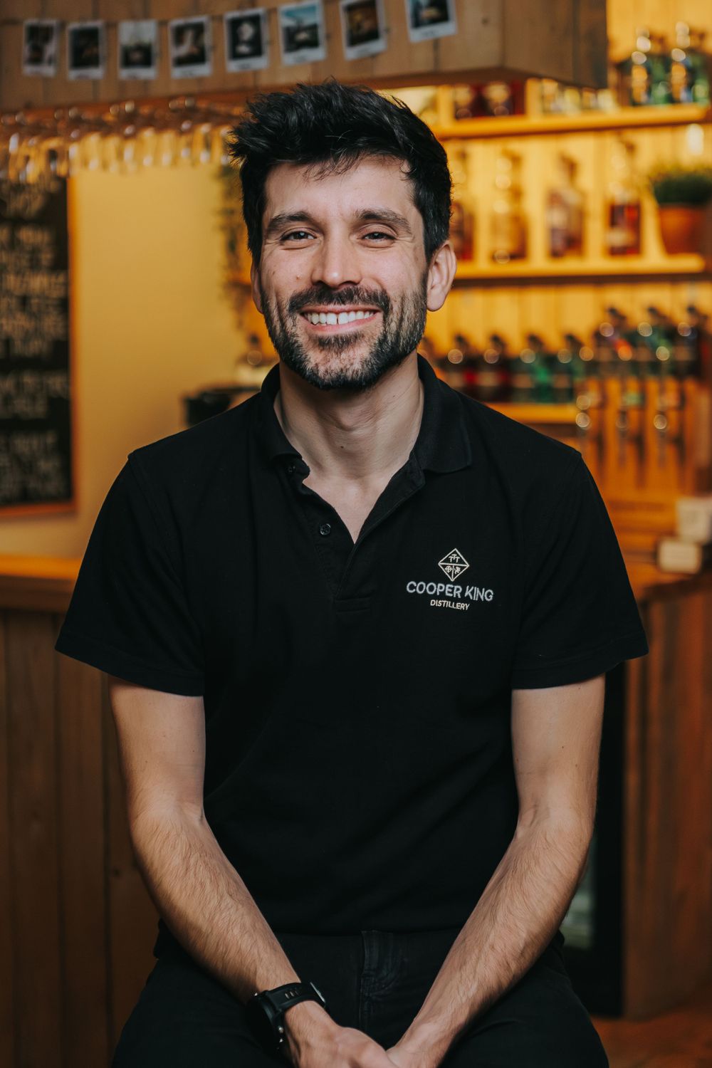 Staff member wearing a black polo shirt with company logo stands in front of bar shelving with bottles.
