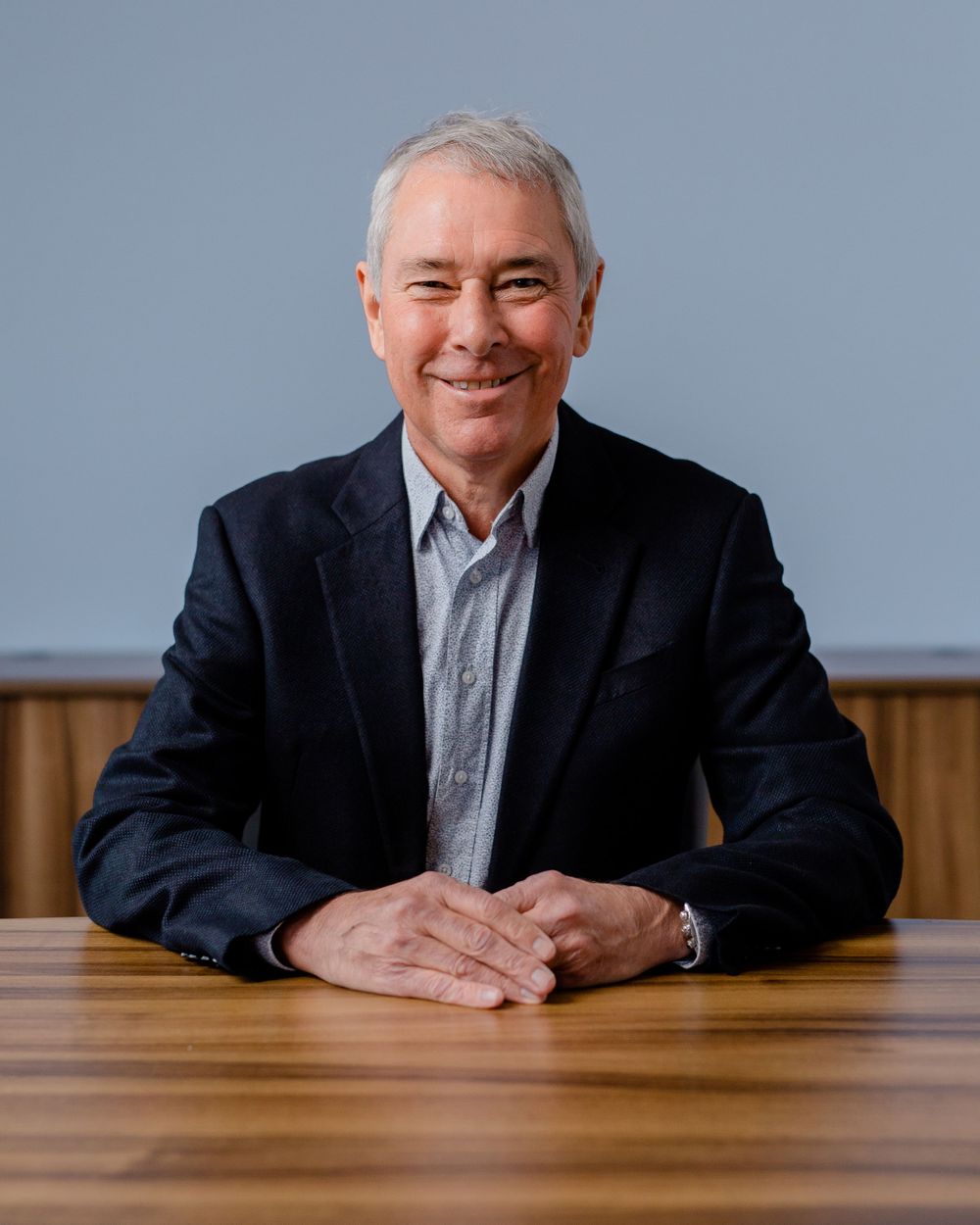 Executive in dark blazer poses professionally at wooden conference table.