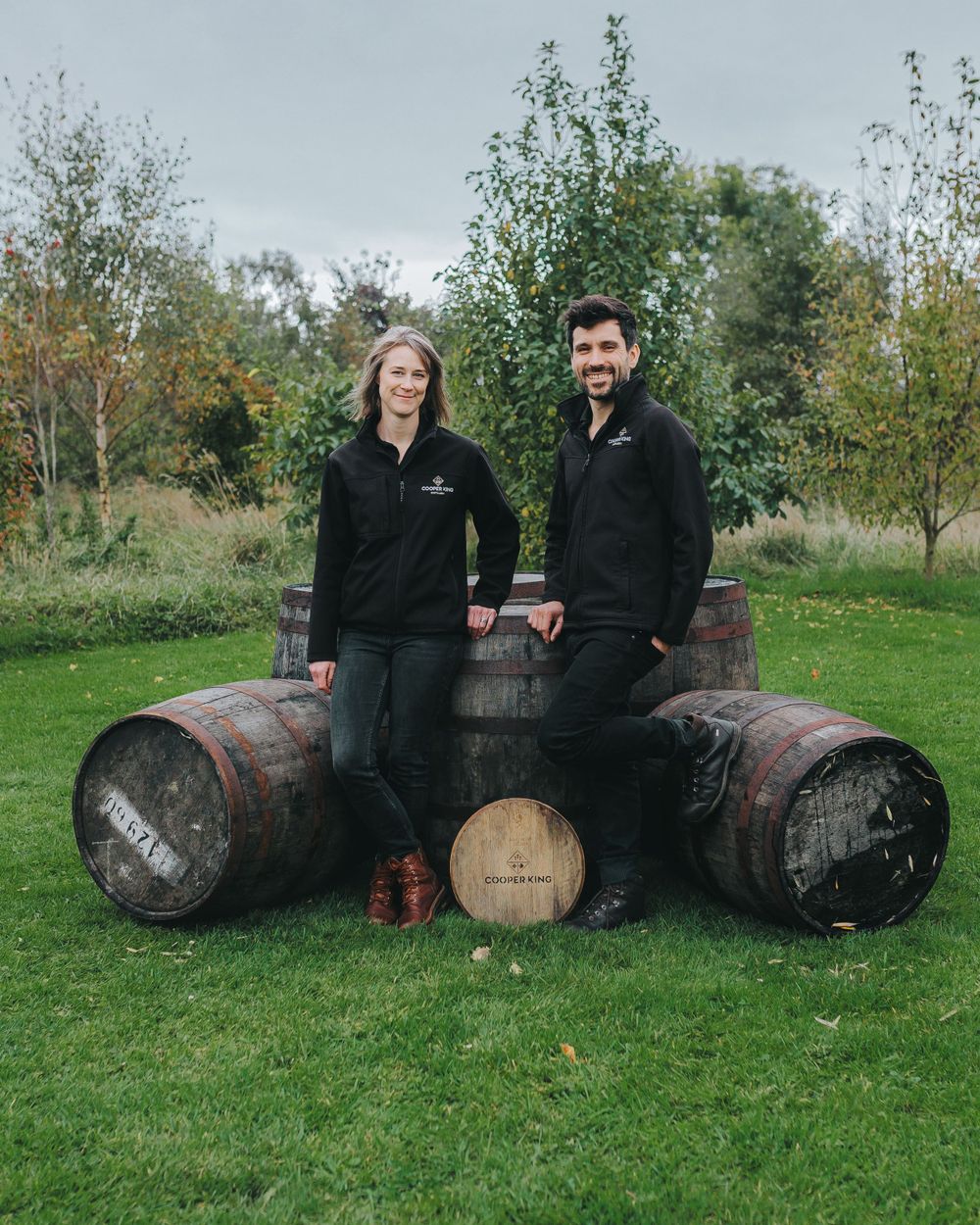 Two employees in matching black uniforms stand beside large wooden barrels in a rustic outdoor setting.
