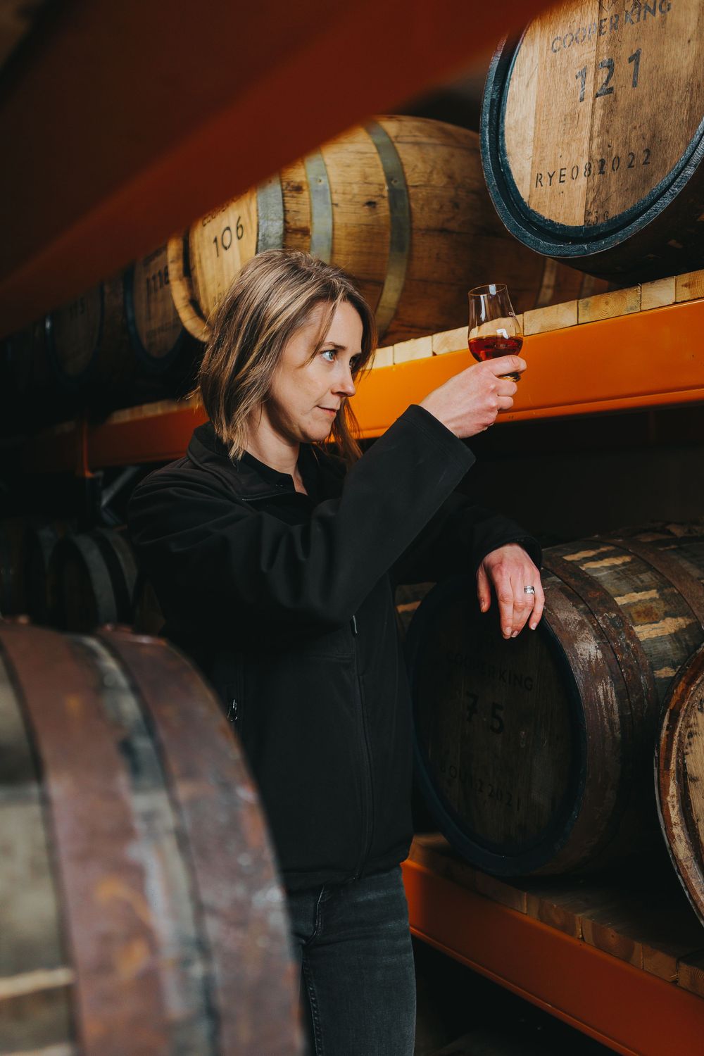 Someone inspects a glass of whiskey in a warehouse filled with aging barrels.