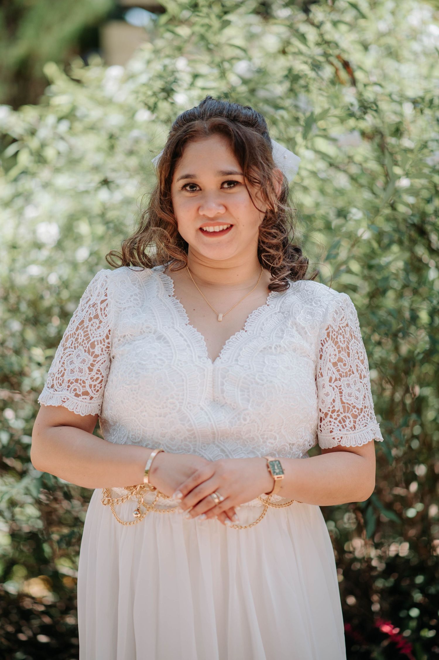 Bride in lace-sleeved wedding dress poses in garden setting with natural lighting.