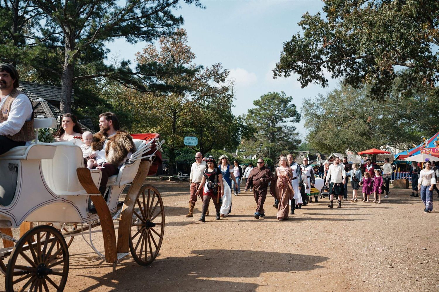 Horse-drawn carriages and festival visitors fill a busy Renaissance fair pathway.