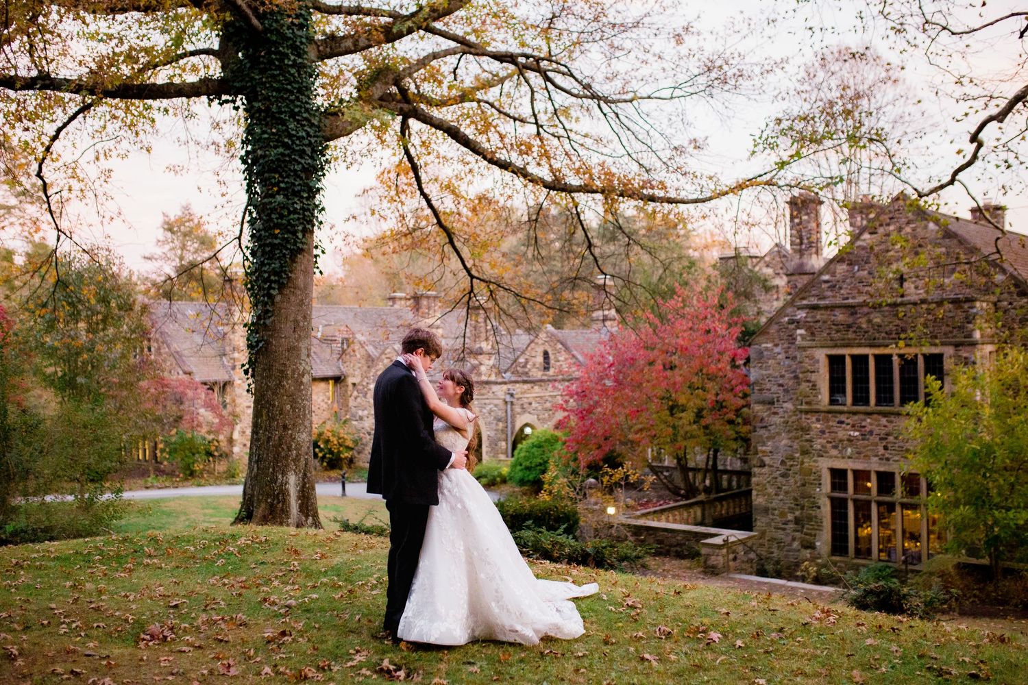 a wedding couple stands with their arms around each other; the sky is pink and there is a stone mansion behind them