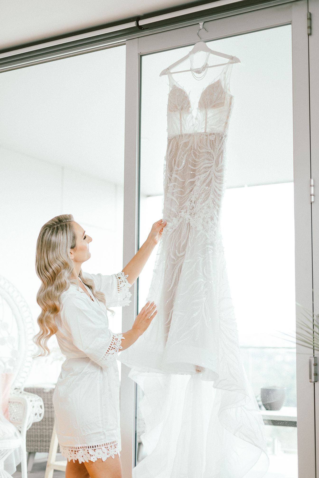 A beautiful white lace wedding dress hangs elegantly by a window as someone admires its intricate details.
