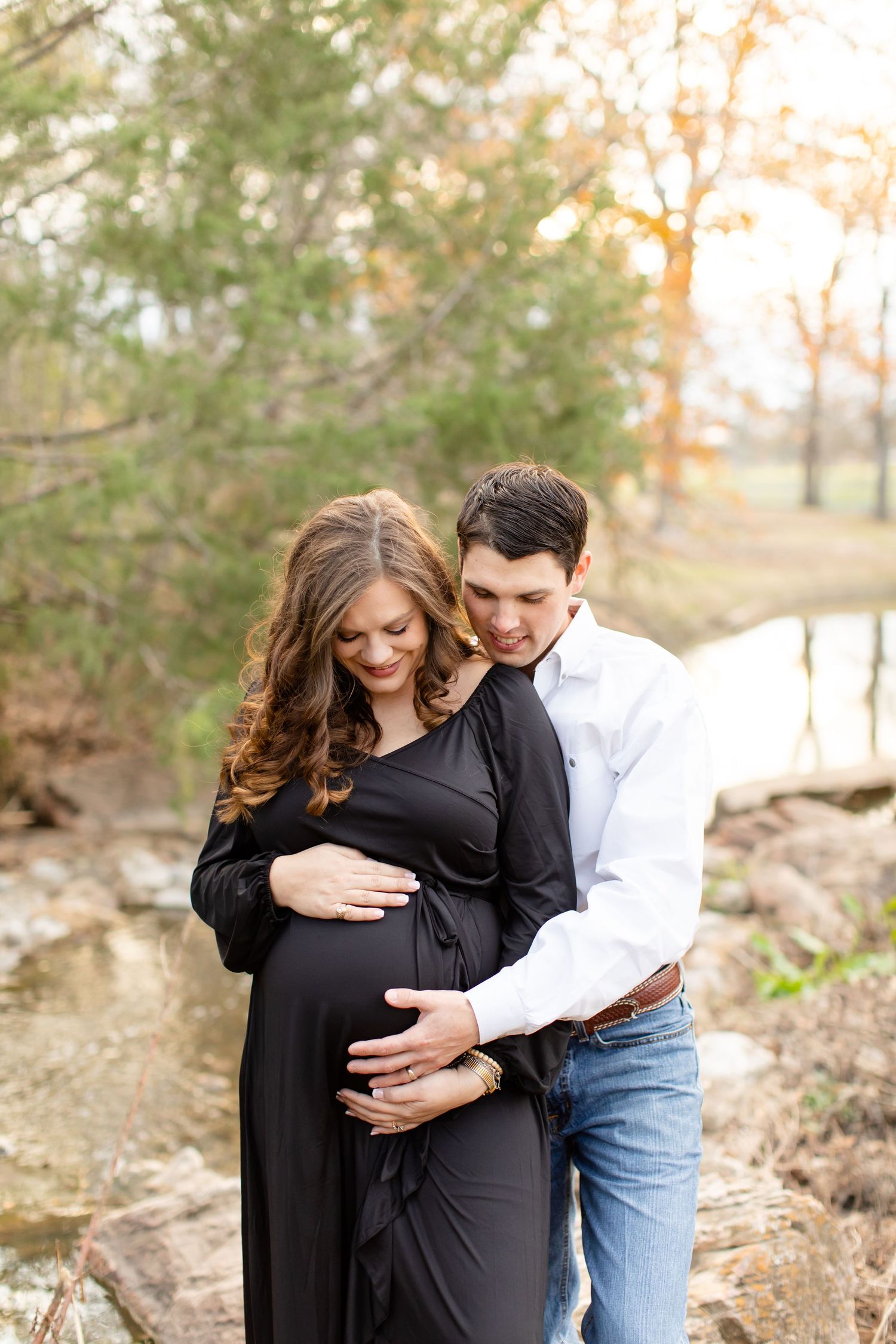Intimate maternity moment captured by water with couple in black dress and white shirt.