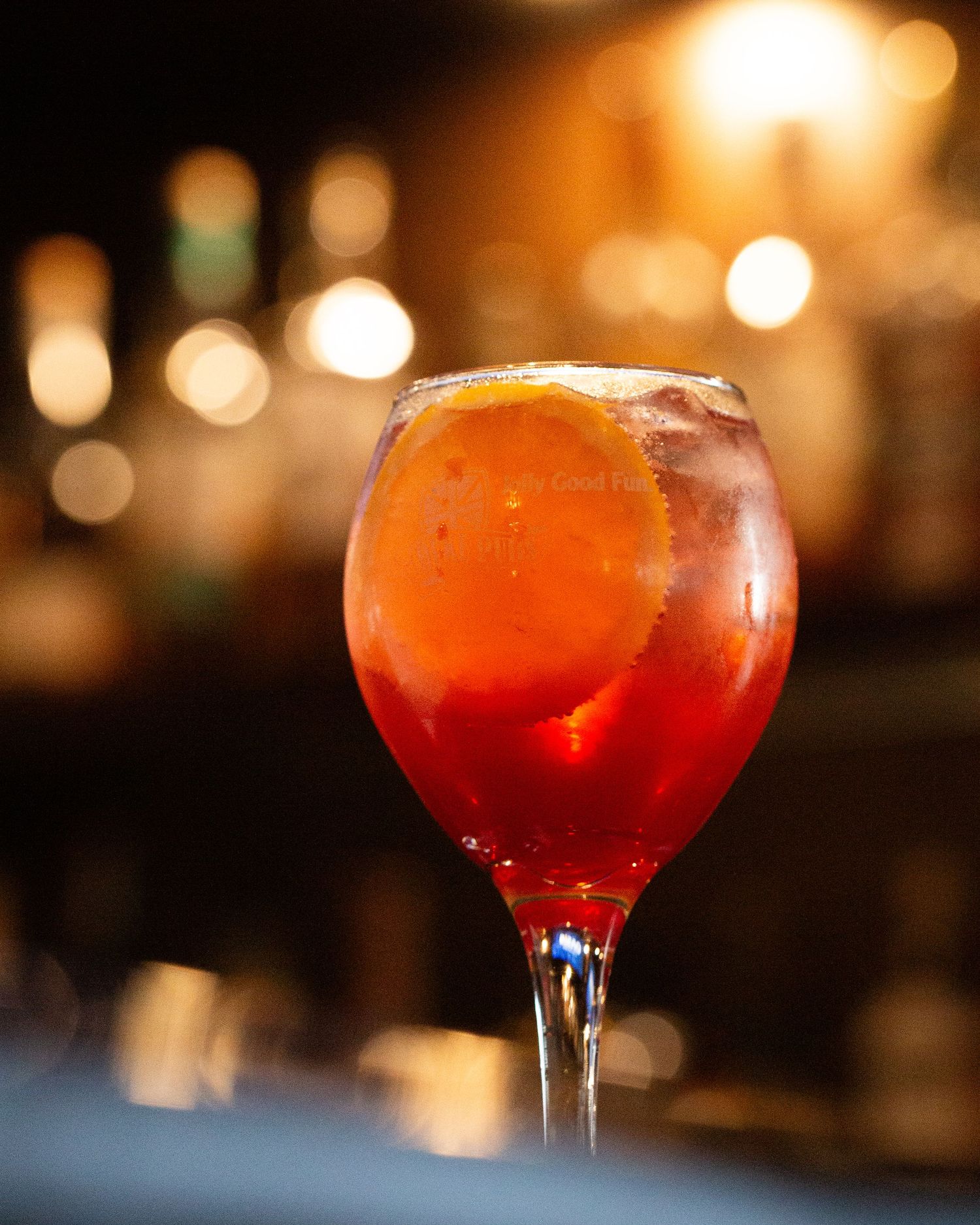 A red cocktail with ice in a wine glass glows warmly against a blurred background with bokeh lights.